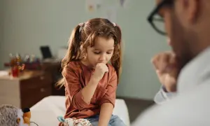 Niña con tos perruna en consulta médica. Un doctor la examina mientras ella se cubre la boca.