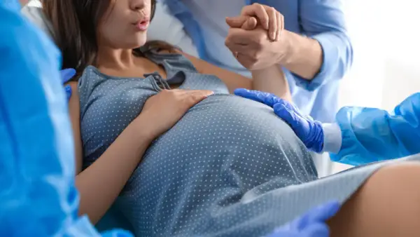 Mujer embarazada en trabajo de parto, con su pareja sosteniendo su mano y personal médico asistiendo. Se enfoca en el vientre, sugiriendo el momento de cómo provocar el parto.